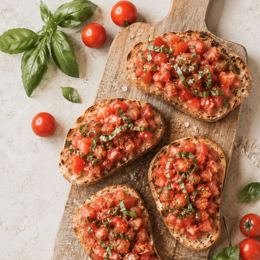 Four slices of toasted bread topped with fresh tomato salsa and basil, arranged on a wooden serving board with whole cherry tomatoes and basil leaves around it on a light stone surface.