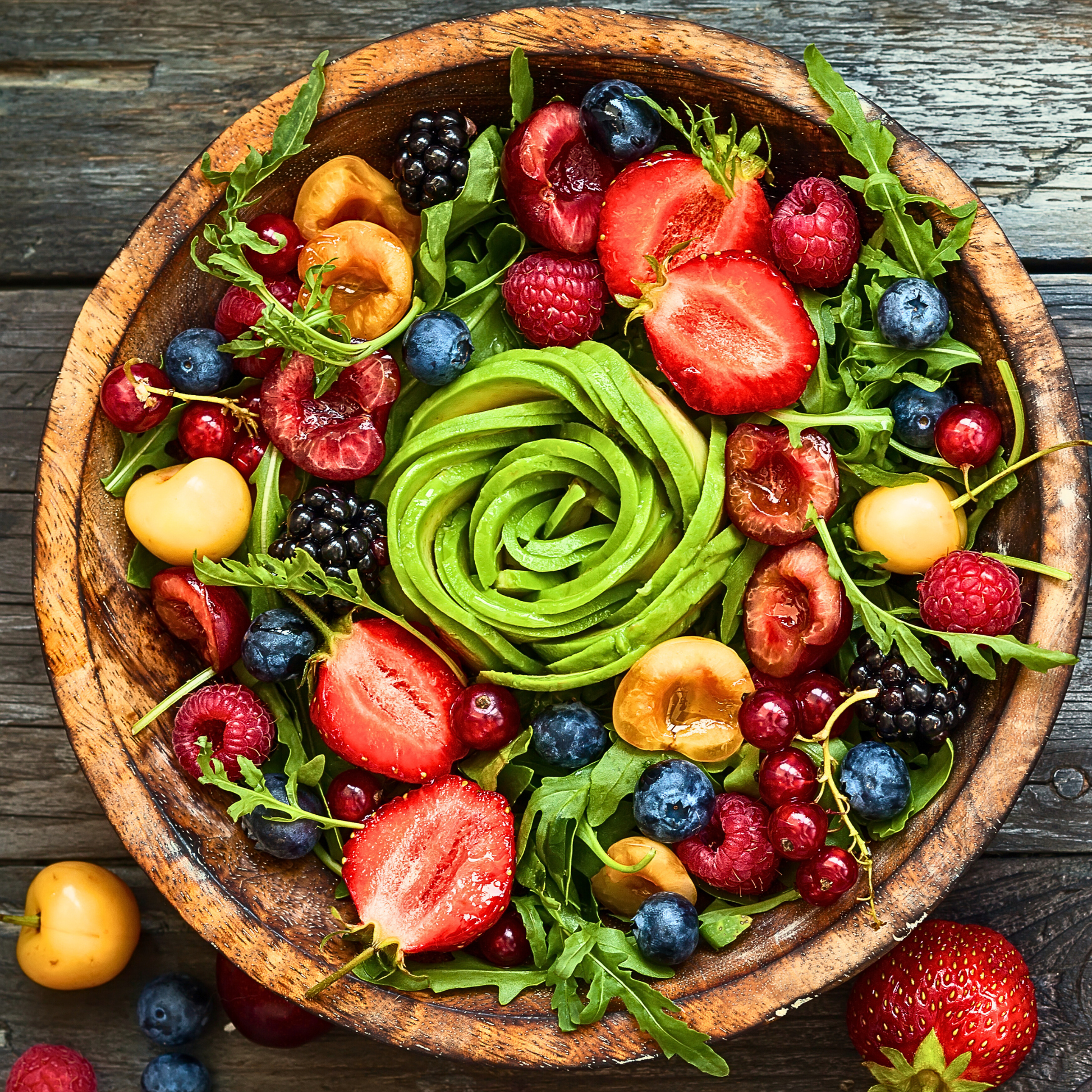 Wooden bowl filled with a colorful salad of fruits, vegetables, and avocados on a wooden surface.