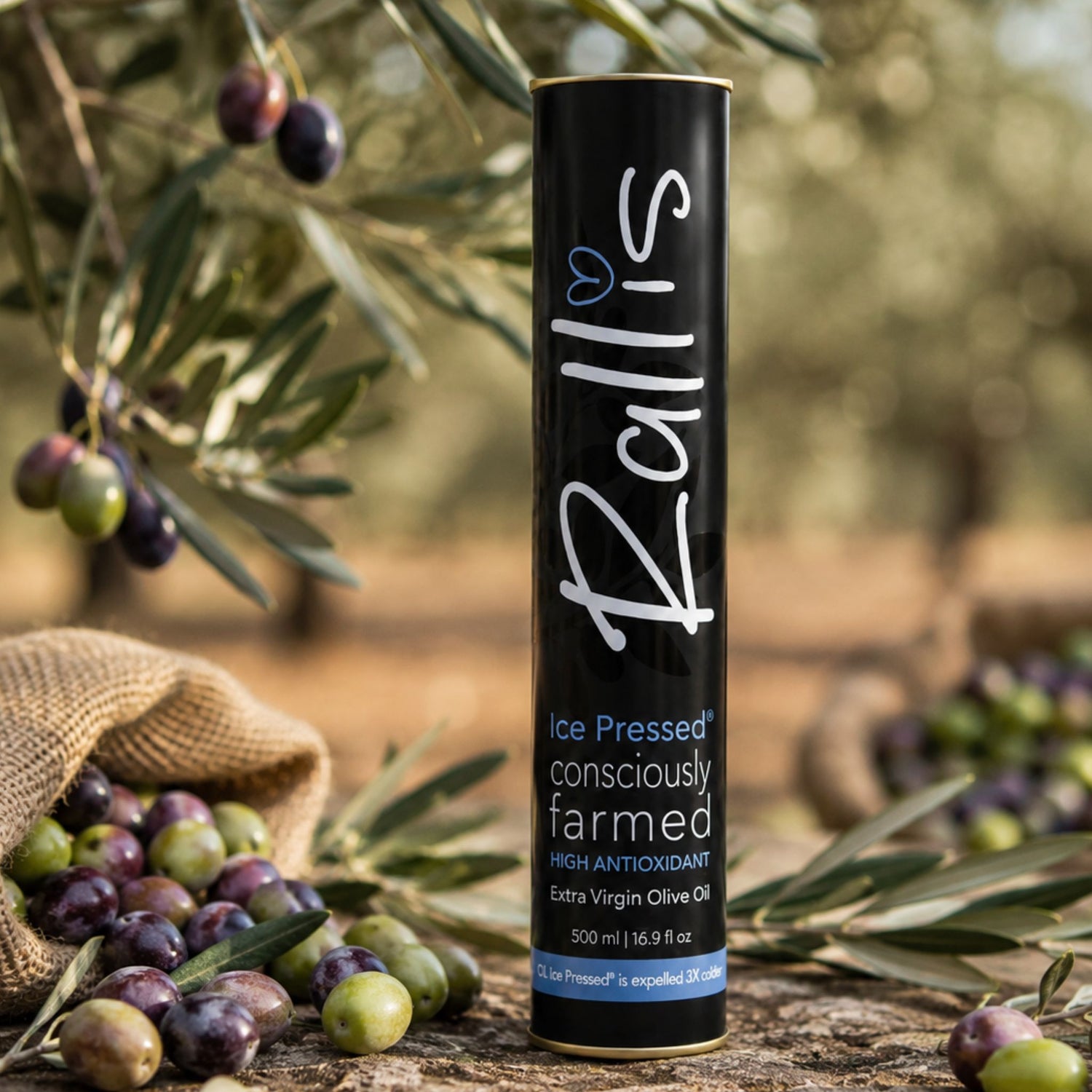 A black cylindrical bottle of Rallis Ice Pressed extra virgin olive oil stands on a wooden surface outdoors, surrounded by olives, olive branches, and a burlap sack. The bottle label is black with white and blue text.