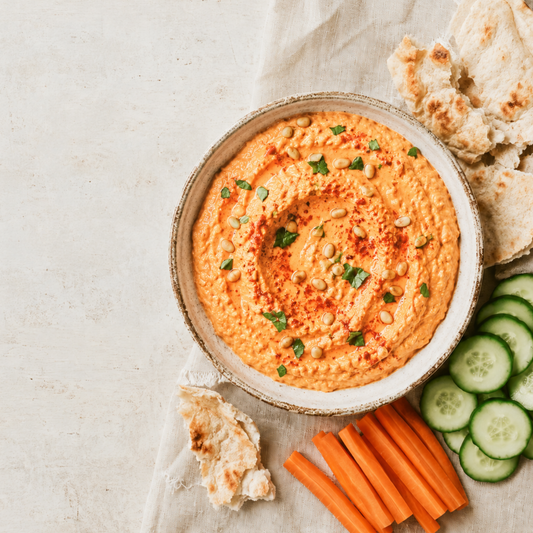 A bowl of thick orange-hued hummus dip with visible herbs, seeds, and red seasoning, served with pita bread, carrot sticks, and cucumber slices on a light neutral surface.