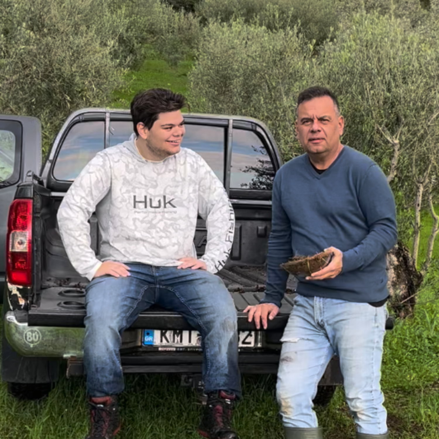 Two men standing outdoors in front of a pickup truck with olive trees in the background. One man is sitting on the tailgate wearing a light gray camouflage hoodie with 'HUK' printed on the chest, and the other is standing wearing a blue long-sleeve shirt and light jeans while holding a dark brown rectangular item.